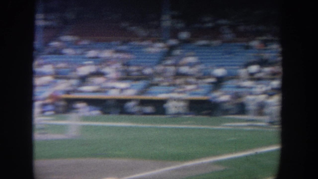 Soft 8mm frame from a New York baseball stadium, looking across the field toward blue seats, spectators, and a dugout area.