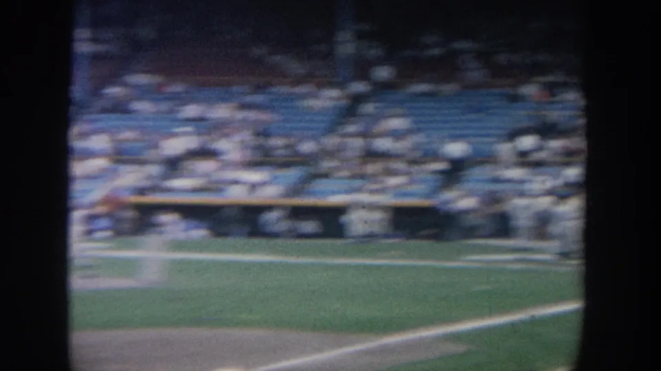 Soft 8mm frame from a New York baseball stadium, looking across the field toward blue seats, spectators, and a dugout area.