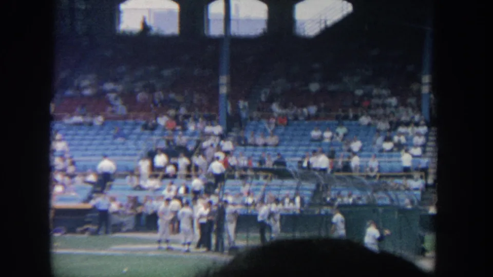 1964 New York stadium frame showing players gathered by the dugout and batting cage, with blue seating and spectators behind them.