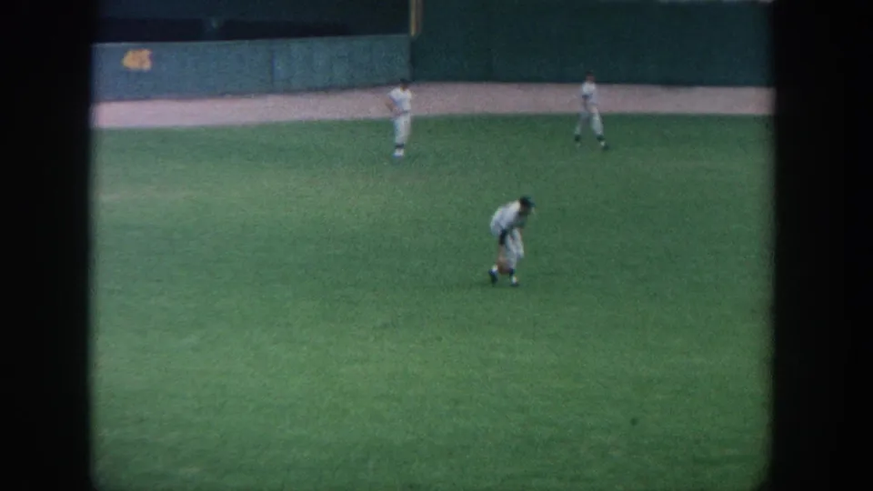New York 1964 home-movie frame showing outfielders spaced across the green field in front of a dark stadium wall.
