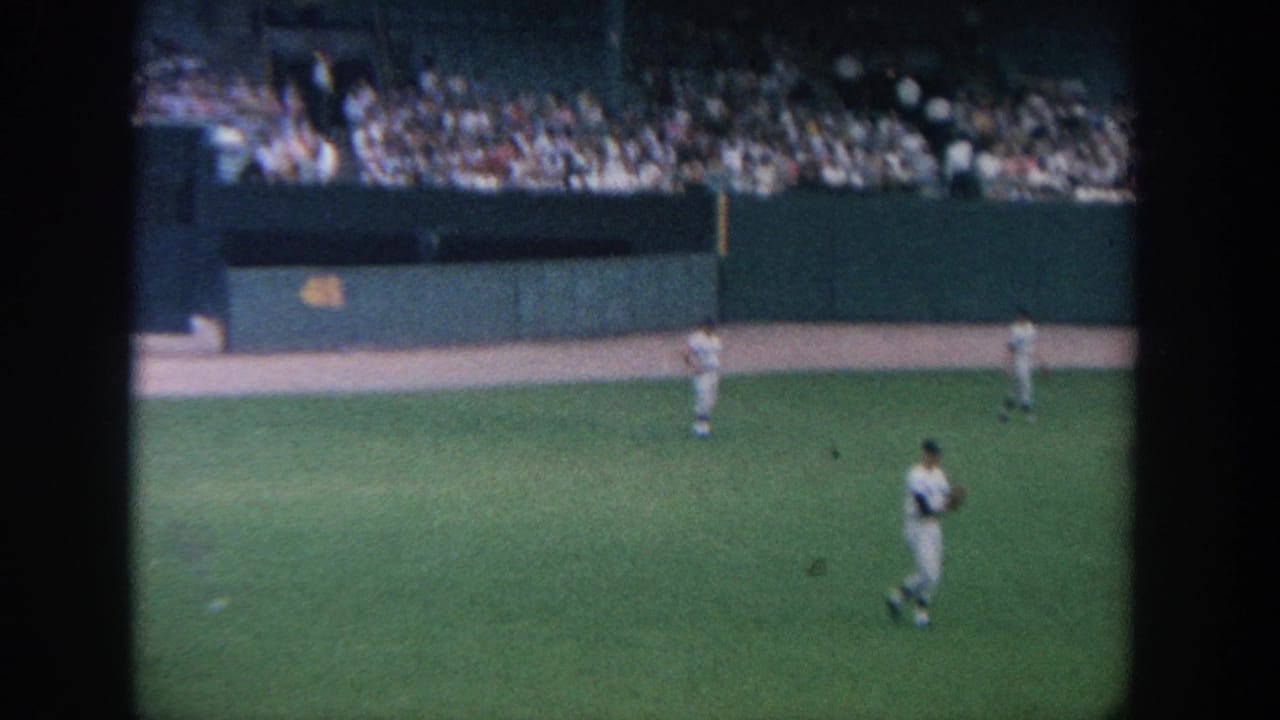 1964 New York baseball game seen from the stands, with white-uniformed players on the outfield grass and spectators in the upper deck.