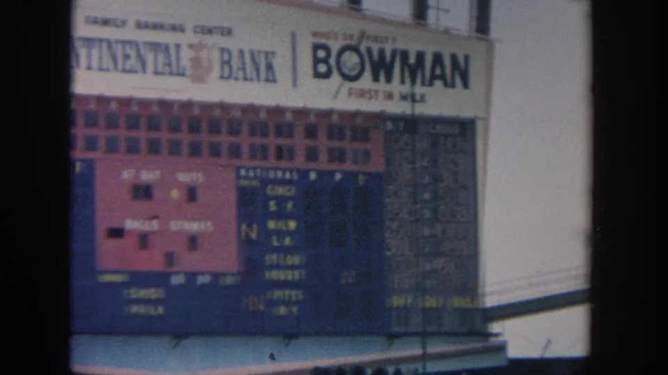 Scoreboard detail from the 1964 New York stadium footage, showing baseball standings information and prominent branded signage.
