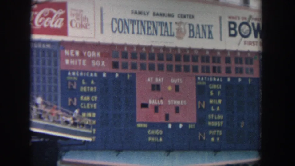 A 1960s New York baseball scoreboard fills the upper frame, with spectators in blue seats below and brand signage along the top.