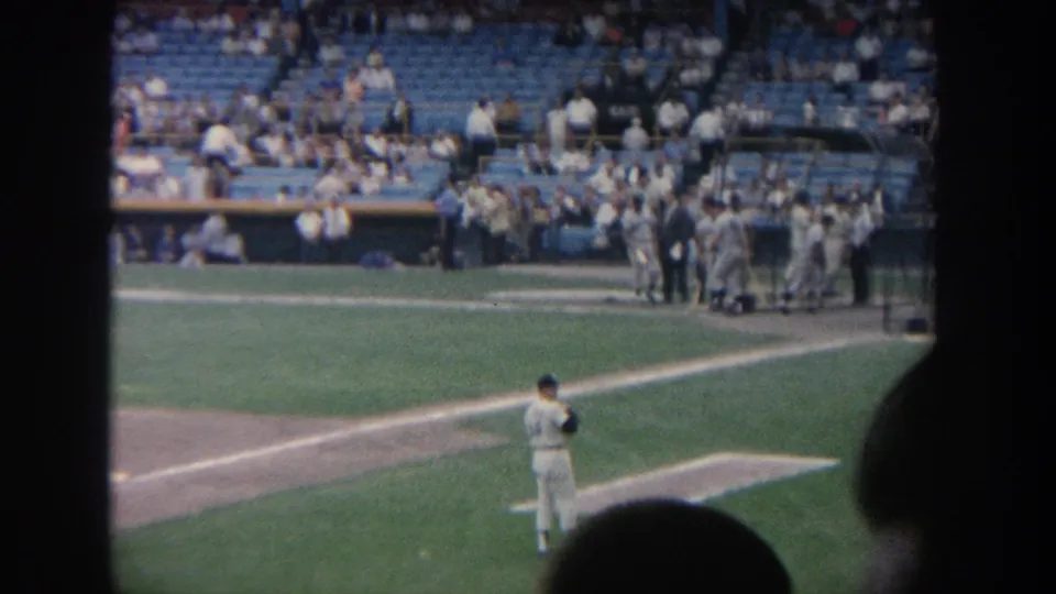 The camera sweep settles on the field, showing a pitcher in white, players near the dugout, spectators, blue seats, and the stadium scoreboard.