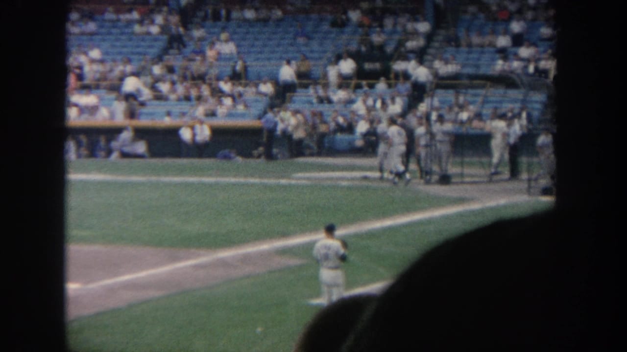 1964 New York home-movie view of a baseball field with pitcher, dugout activity, blue stadium seating, and dark spectator silhouettes.