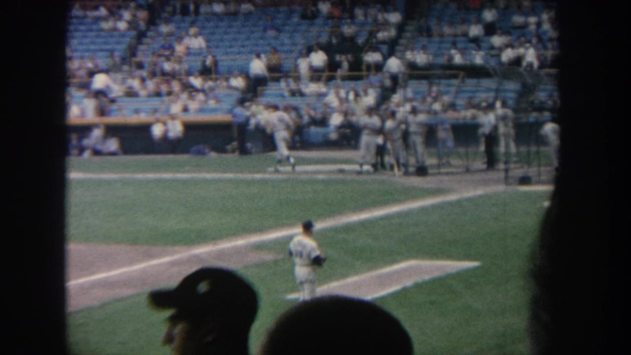 1964 New York baseball game footage showing the pitcher, dugout group, sunlit grass, blue seating, and spectator silhouettes in front.