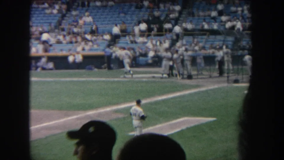 1964 New York baseball game footage showing the pitcher, dugout group, sunlit grass, blue seating, and spectator silhouettes in front.