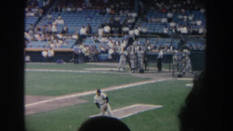 1964 New York home-movie frame with a player in pitching motion, dugout figures, blue seats, and partial spectators in the foreground.