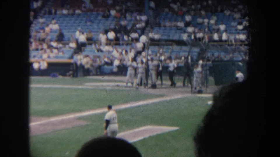 1964 New York spectator-shot baseball footage with pitcher in foreground, dugout group, crowd seating, and dark silhouettes at frame edges.