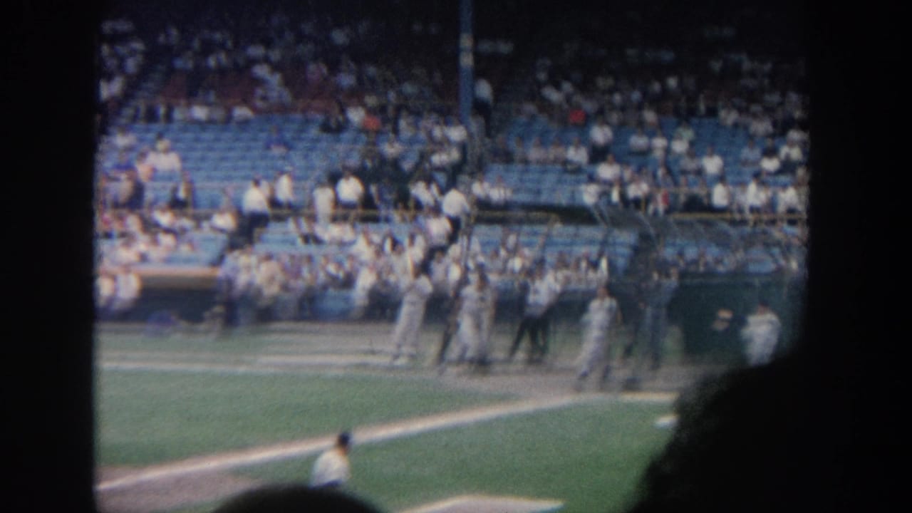 1964 New York 8mm view of a baseball game, with players near the infield and stadium spectators visible through dark foreground edges.