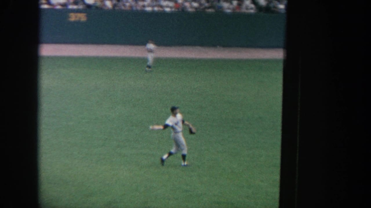 Outfield player in a white uniform moving across green grass with another player in the distance.