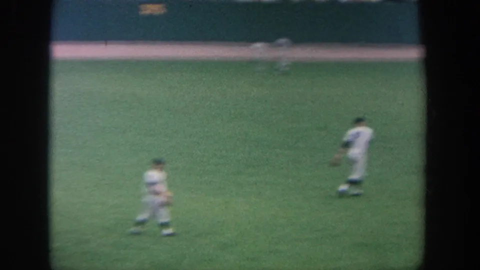 Two players in white uniforms are visible on the outfield grass in soft-focus 8mm footage from a 1964 New York baseball game.