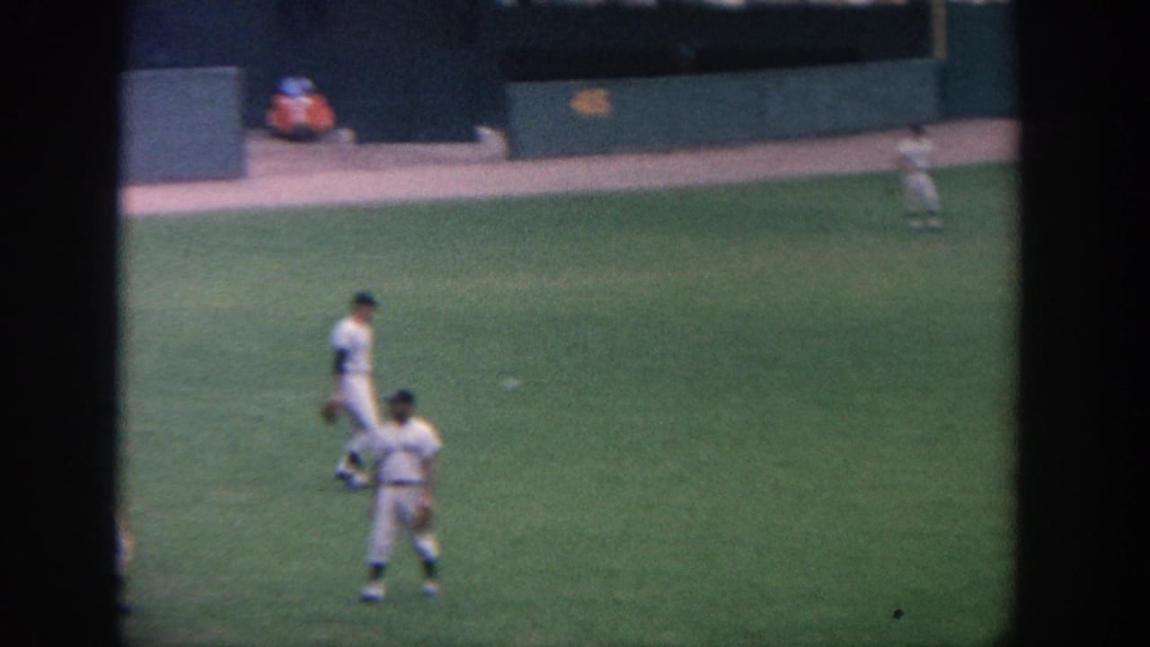 Baseball players in white uniforms stand on green outfield grass near a dark stadium wall.