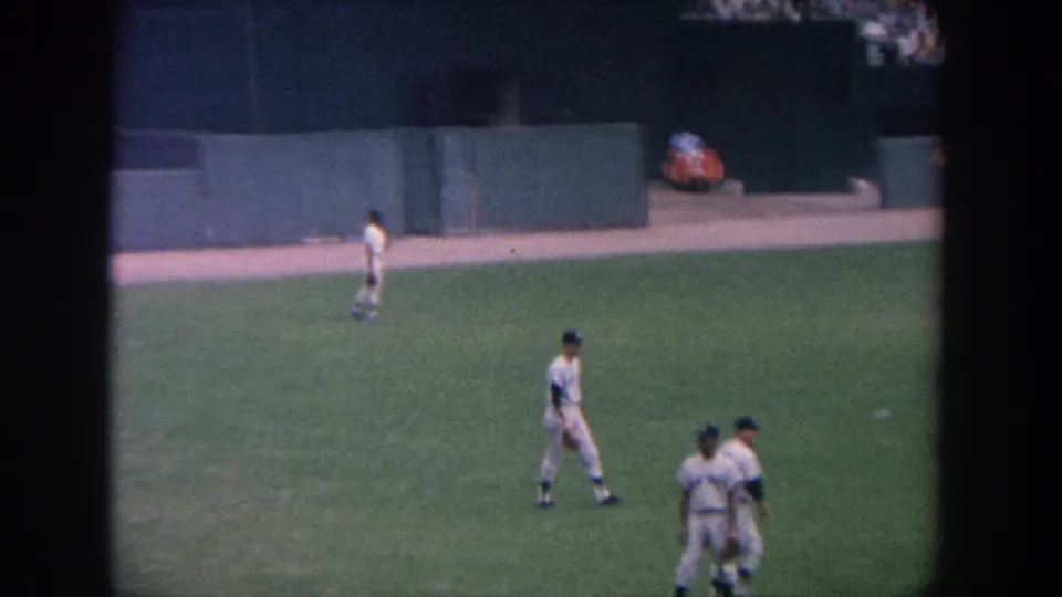 Players in white uniforms move across the grass during a 1964 New York baseball game, filmed from the stands.