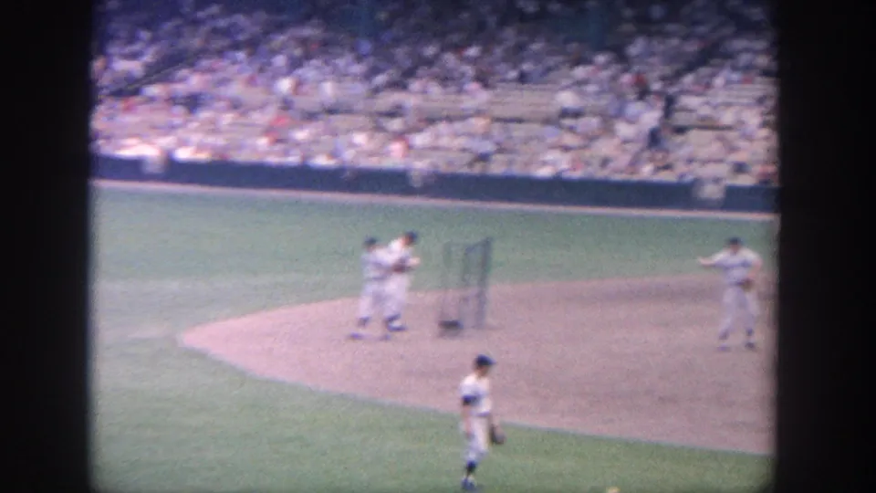 A home-movie view of the infield shows players positioned on dirt and grass with a crowd seated behind them.