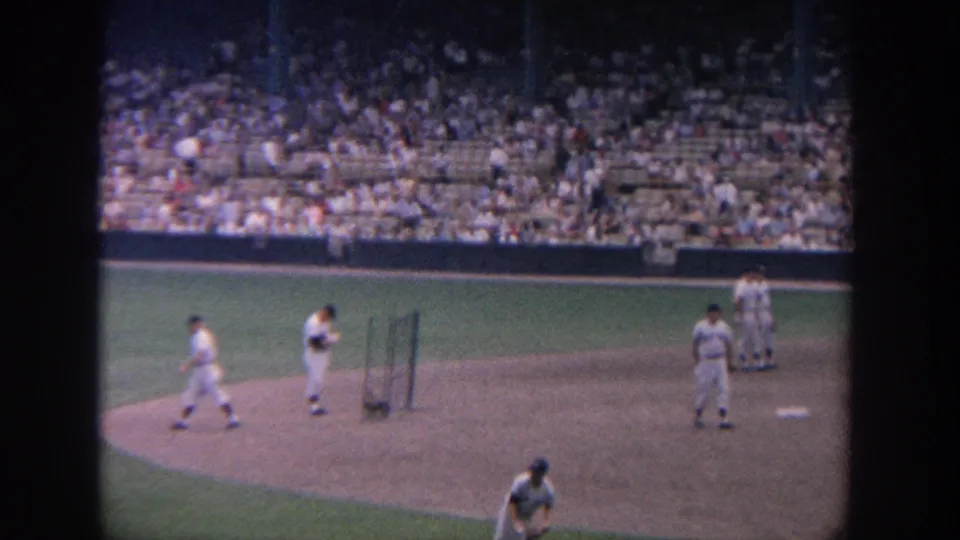The frame shows players around the infield and baseline with packed stadium seating in the background.