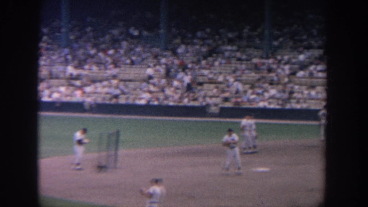 Baseball players stand on a dirt infield with a crowded stadium section behind them.