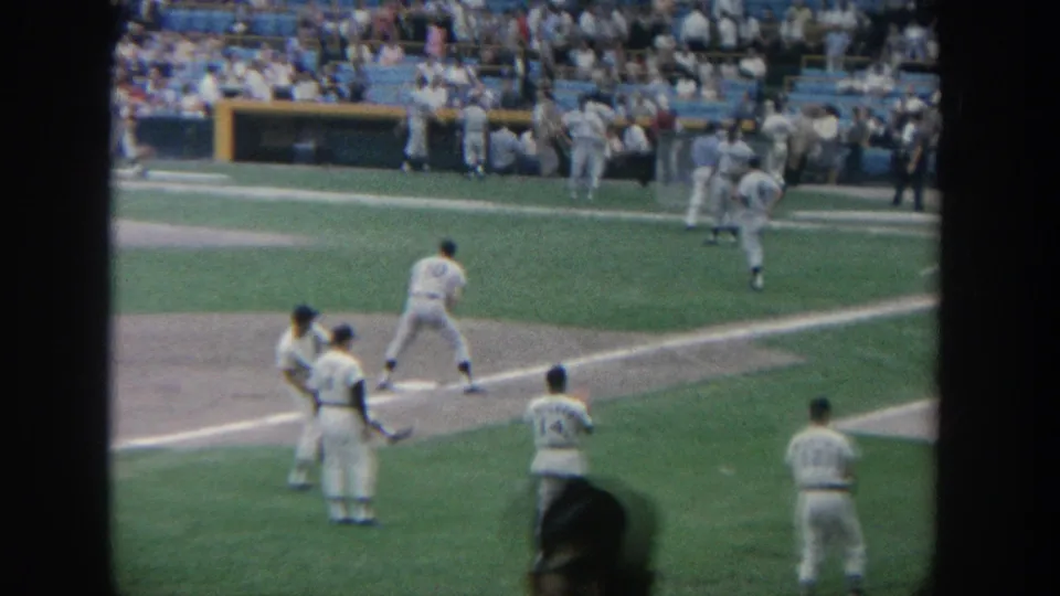 1964 New York 8mm frame showing baseball players near a base line, with dugout area, field grass, and spectators beyond.