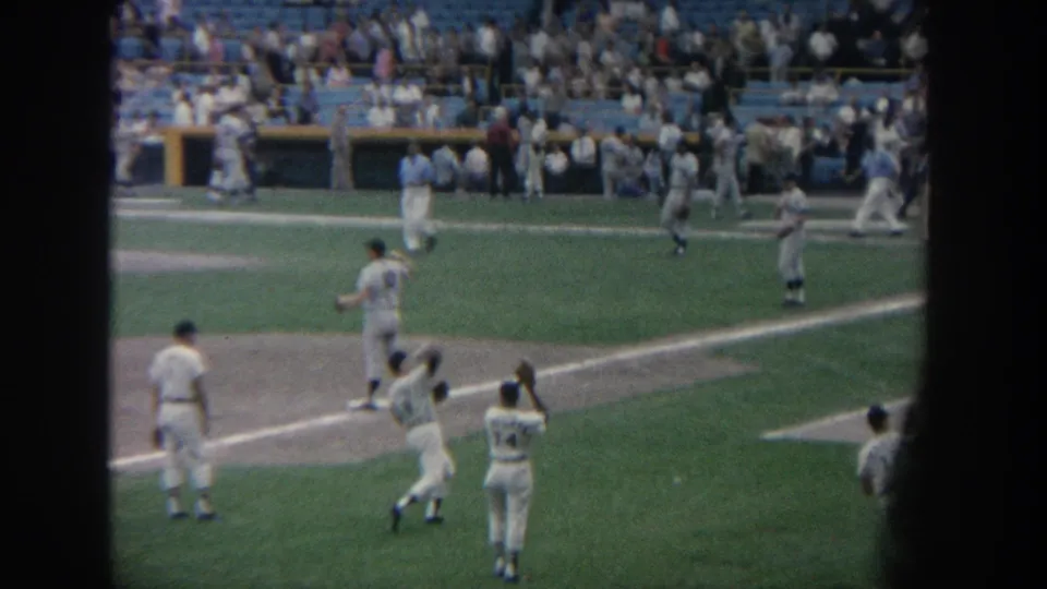 1964 New York 8mm frame showing a baseball player reaching with a glove as teammates warm up before a game.