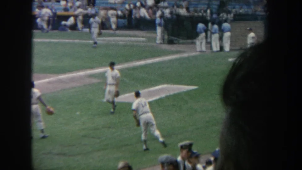 A 1964 New York ballpark scene shows players on the green field, blue seating, spectators, and dark home-movie framing at the edge.