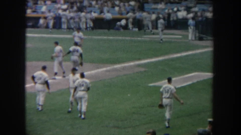 A 1960s ballpark view shows players in white uniforms walking and warming up near the infield with spectators behind them.
