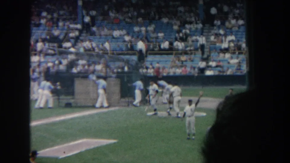 A wider 1964 New York ballpark view shows players near the dugout, a player in the foreground, and spectators filling the seats.