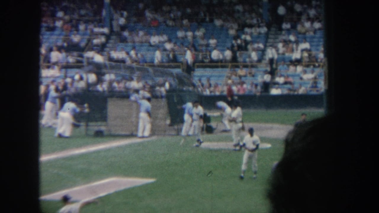 1964 New York home-movie view of a baseball field with players near the infield and fans in blue stadium seats.