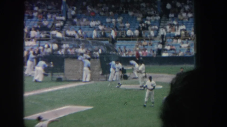 1964 New York home-movie view of a baseball field with players near the infield and fans in blue stadium seats.