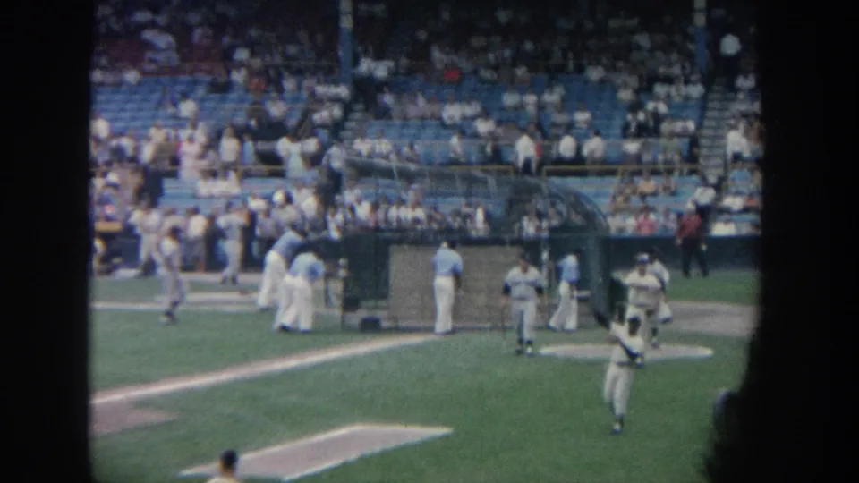 Spectator-shot 1964 New York baseball scene with field activity, a batting area, and distant fans in blue seats.