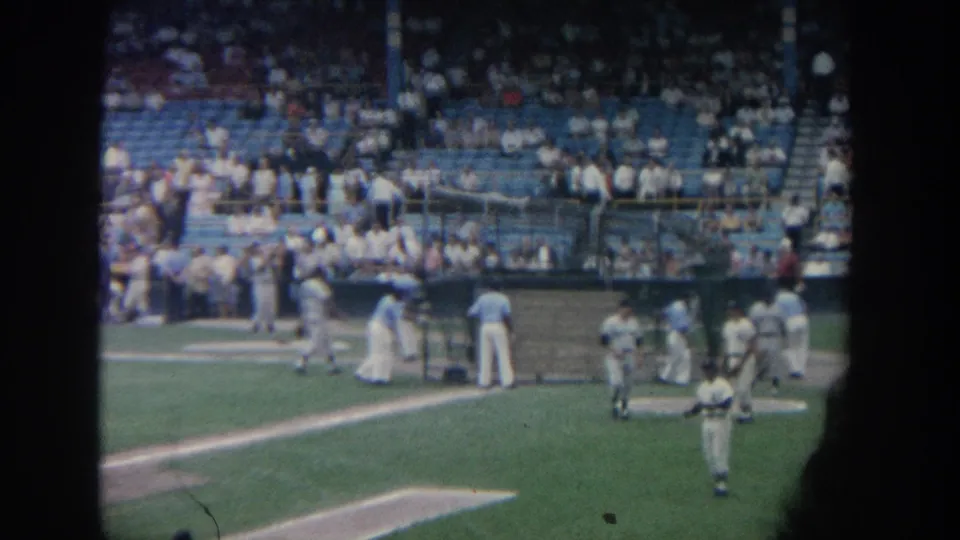 Home-movie frame from a 1964 New York baseball game, showing players near the batting area and fans in the stands.