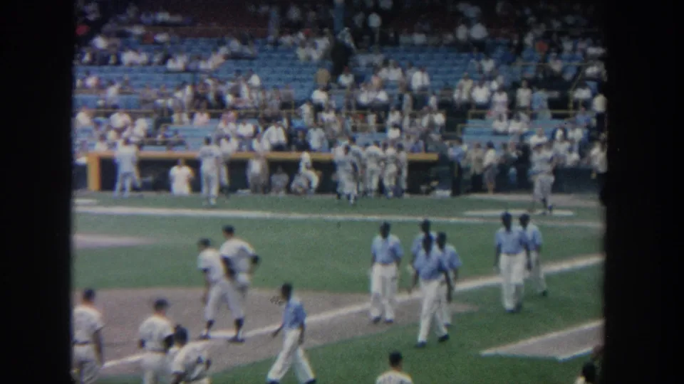 A distant 1964 New York stadium view shows baseball players moving near the infield with a crowd in the stands.