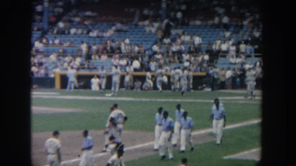 The home-movie frame shows players crossing a New York baseball field, with spectators and dugout activity in the background.
