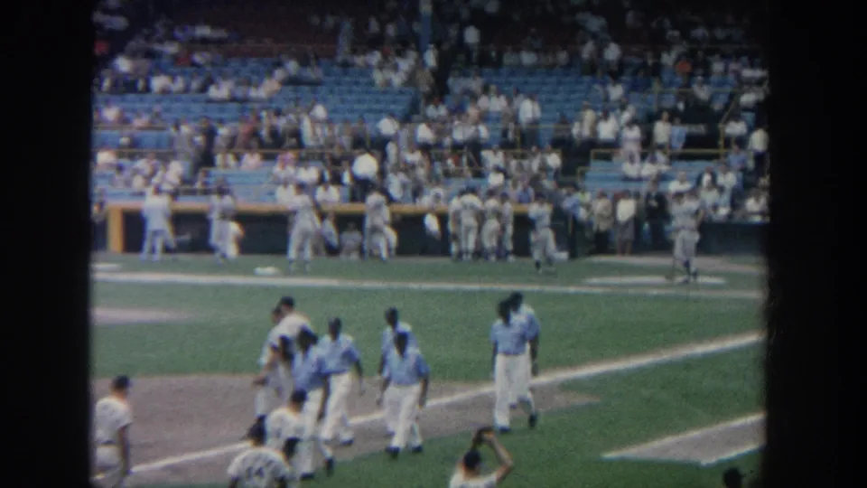 A 1964 New York 8mm frame shows baseball players leaving the infield area while spectators watch from blue seats.