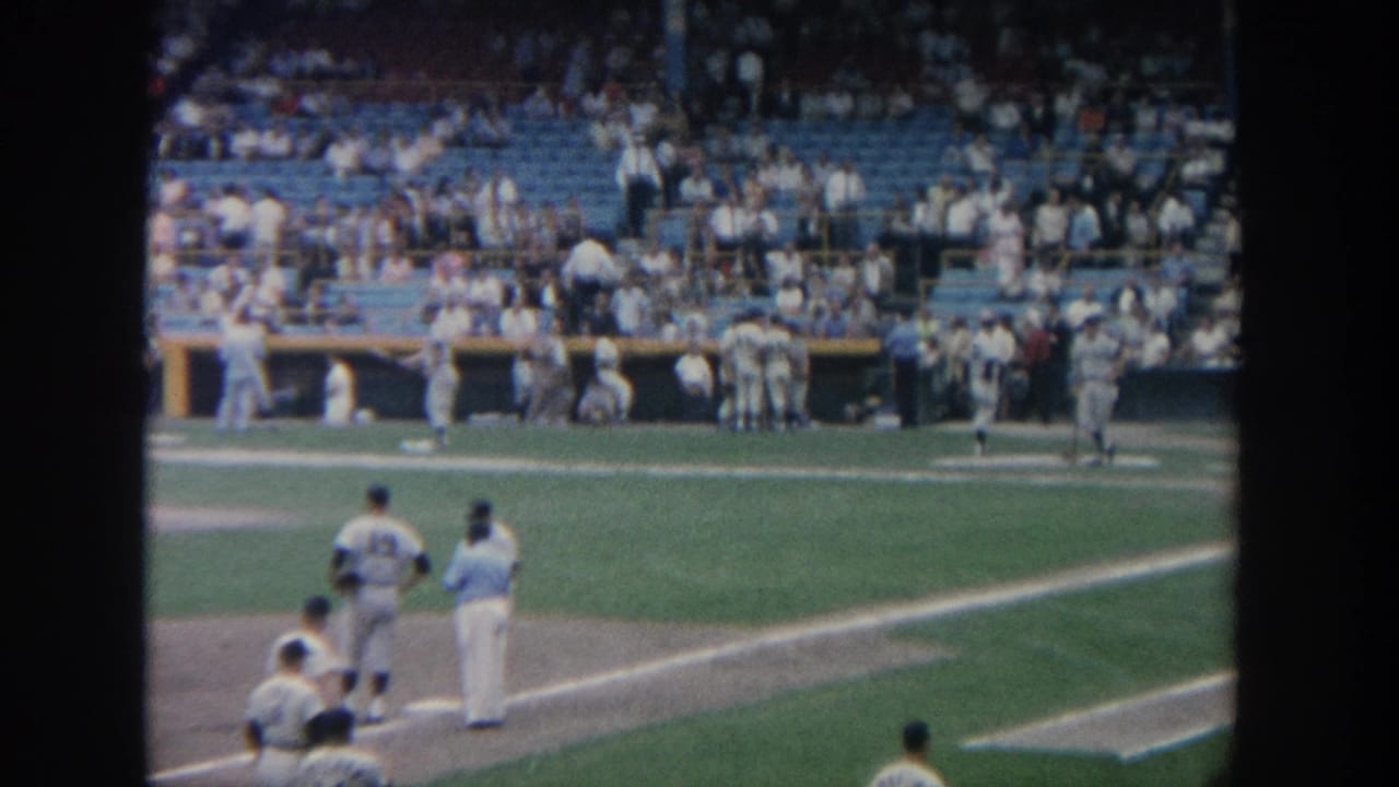 1964 New York baseball stadium view with players in white uniforms near the baseline and fans in blue seats.