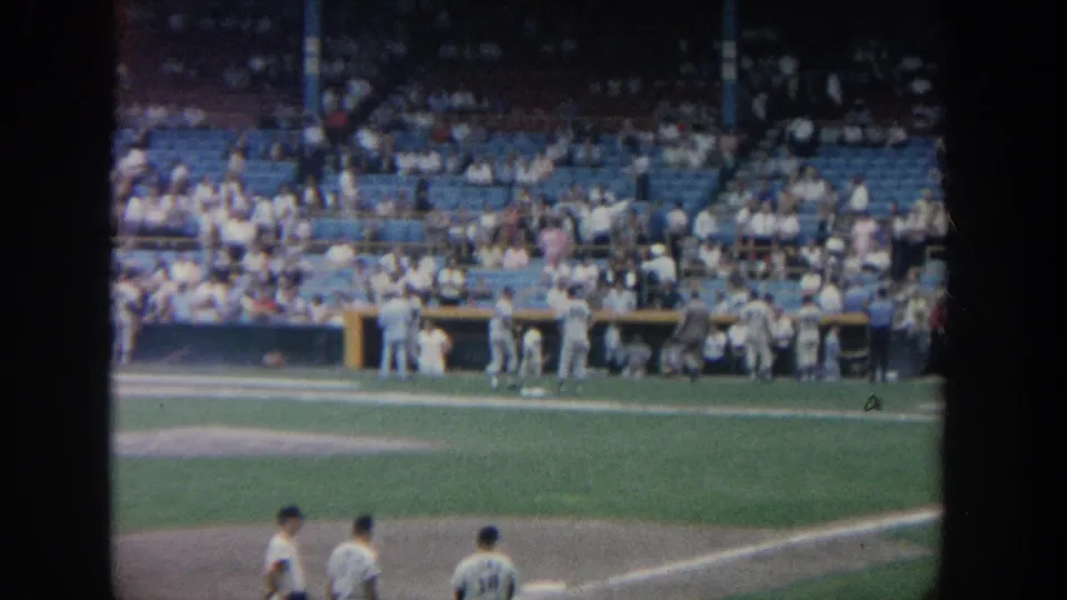 The 8mm frame shows players on the infield and near the dugout, with a spectator crowd behind them.