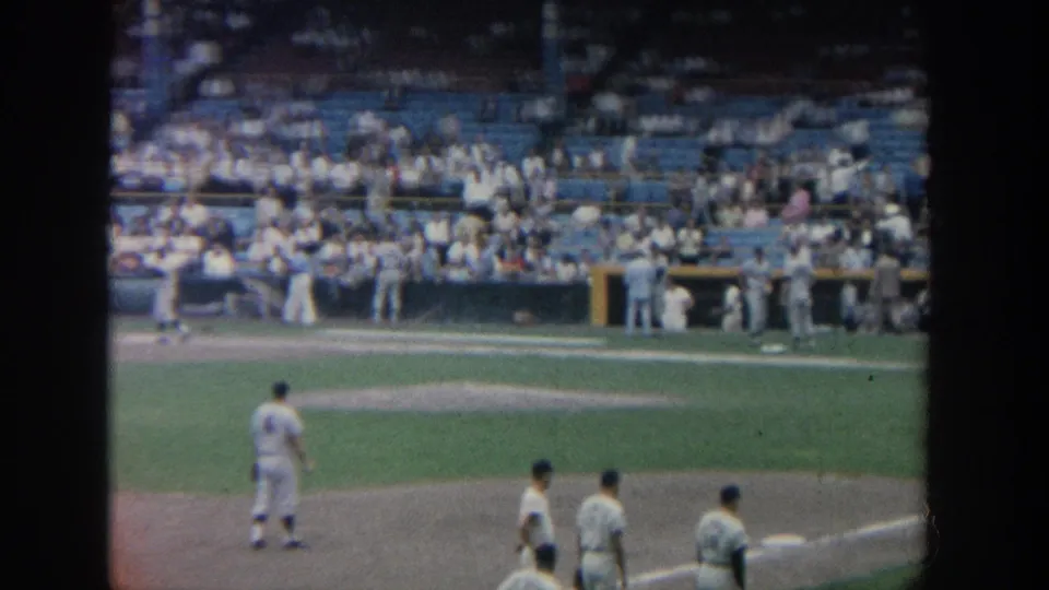 Several players in white uniforms stand near the infield base area in 1964 New York stadium footage.
