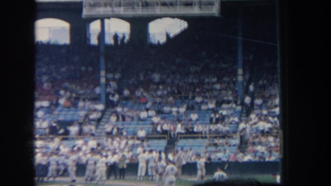 1964 New York home-movie frame showing baseball players in white uniforms near the field edge with spectators filling the stands.