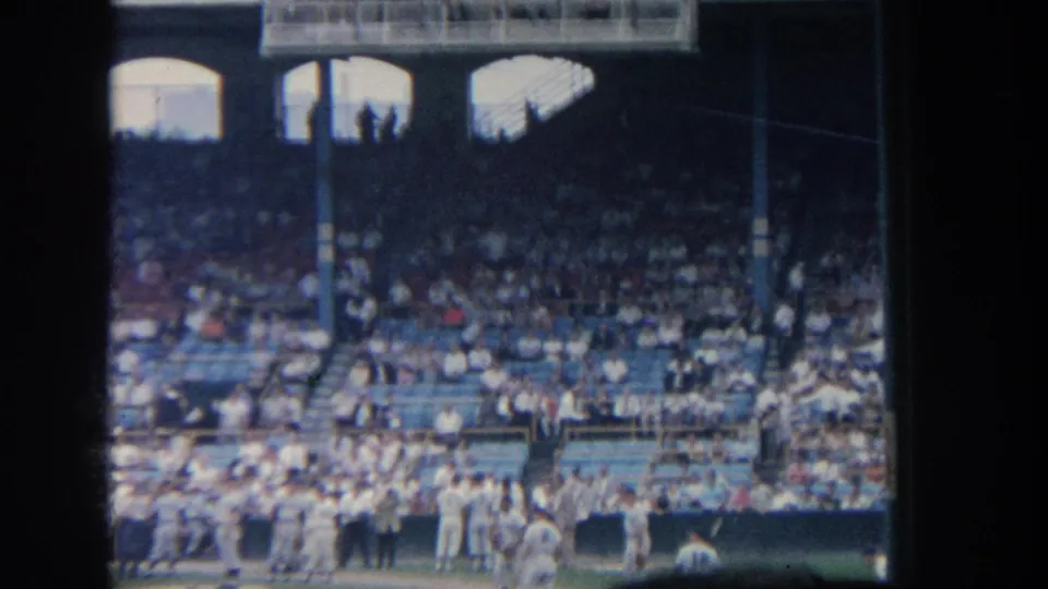 1964 New York home-movie frame showing baseball players in white uniforms near the field edge with spectators filling the stands.