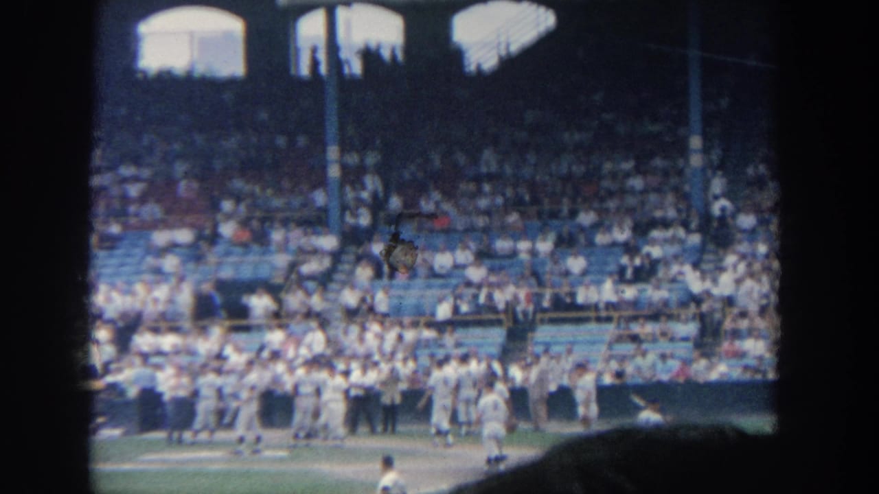 Baseball players and staff stand near the dugout area before a crowded grandstand.
