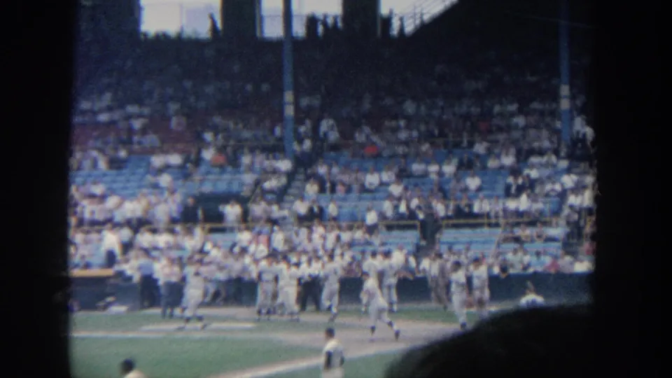 1964 New York 8mm frame showing a baseball team spread along the field edge with spectators seated in blue and white stands.