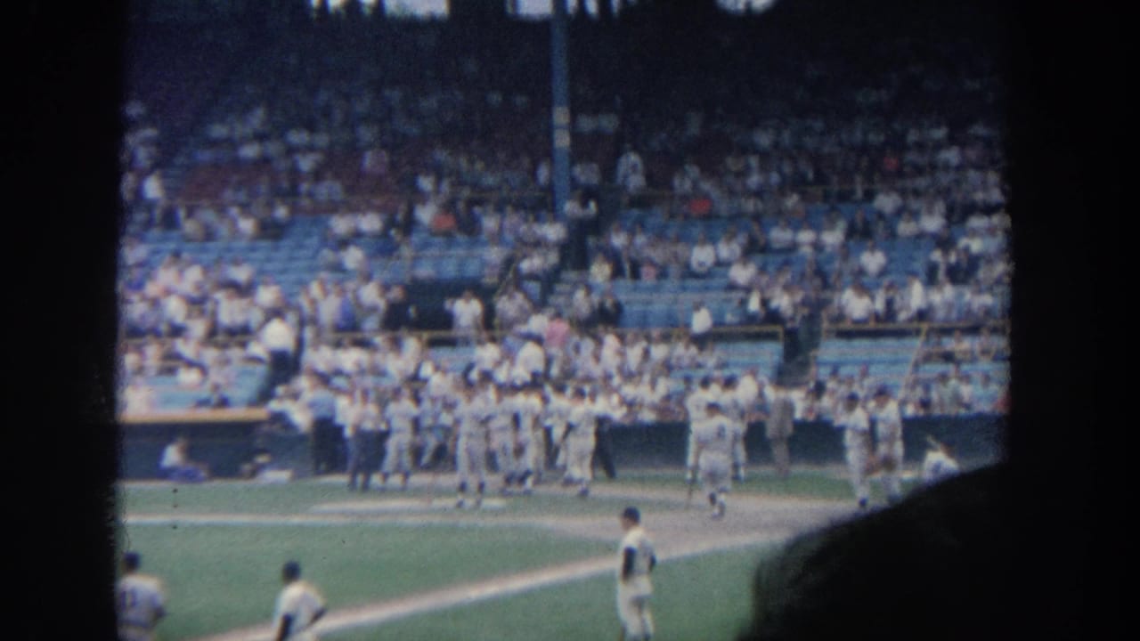1964 New York 8mm frame showing players in white uniforms near a dugout with a crowded stadium behind them.