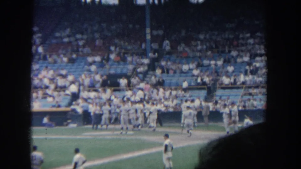 1964 New York 8mm frame showing players in white uniforms near a dugout with a crowded stadium behind them.