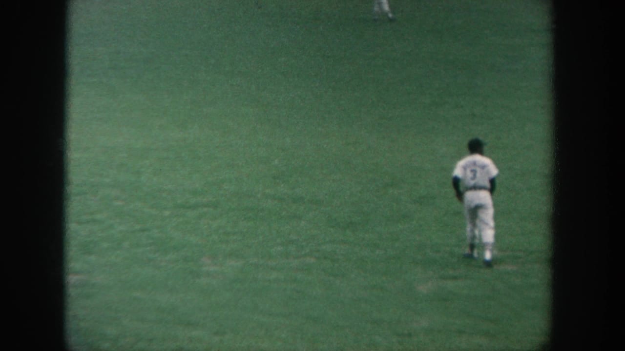 1964 New York 8mm frame showing a baseball player in a white uniform with number 3 standing on the outfield grass.