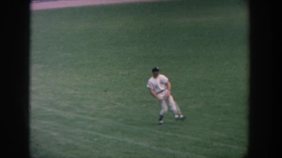 1964 New York 8mm frame showing a uniformed baseball player leaning forward on the green outfield.
