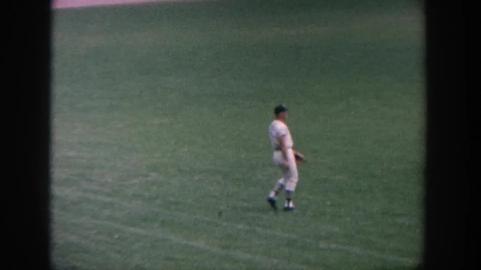 1964 New York home-movie frame of a baseball player in a white uniform standing in the outfield with a glove.