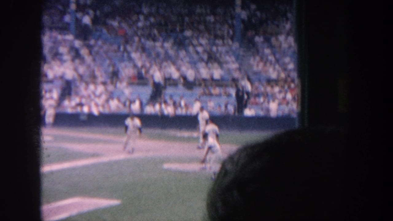 1964 New York baseball game seen from the stands, with players on the field, packed blue seating, and a foreground spectator silhouette.