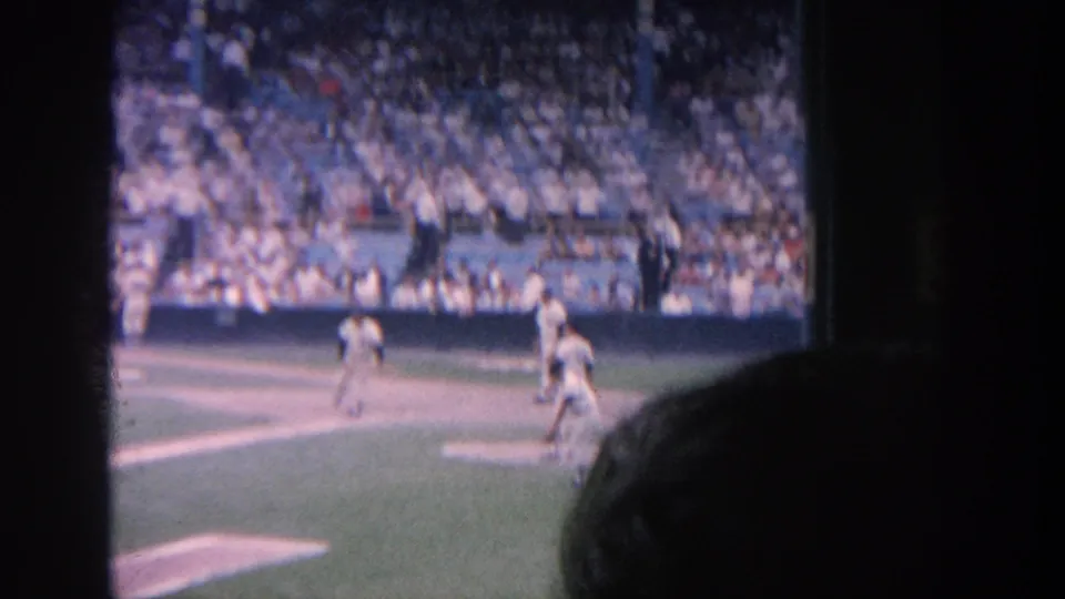 1964 New York baseball game seen from the stands, with players on the field, packed blue seating, and a foreground spectator silhouette.