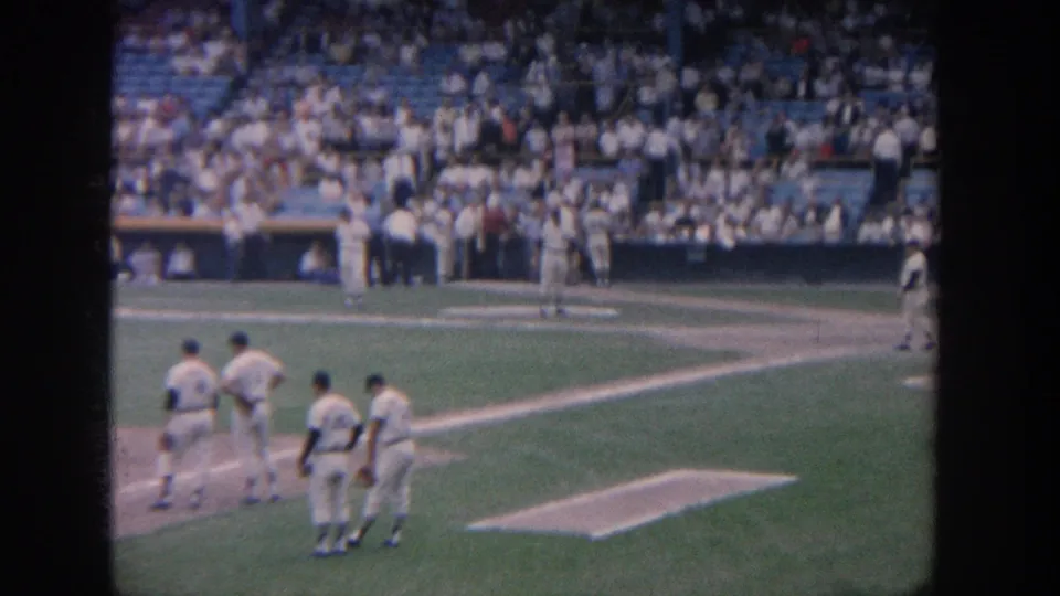 Players in white uniforms gather near the infield during a 1964 New York baseball game, with crowd seating and a dugout visible behind.