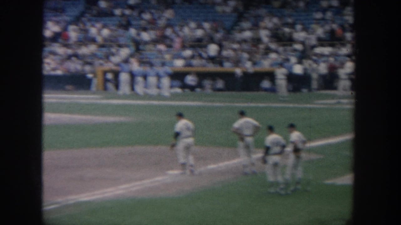 A baseball player moves near first base as several teammates stand nearby and spectators sit in the stands.