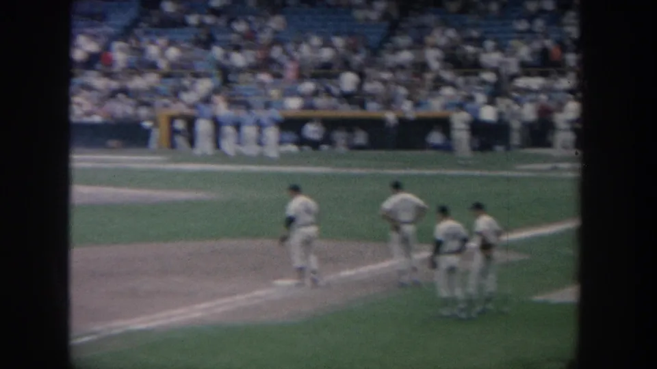 In the 1964 New York baseball scene, players in white uniforms remain near first base with the crowd visible beyond the field.